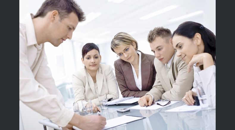 Businessman leaning on desk, explaining to four colleagues sitting.